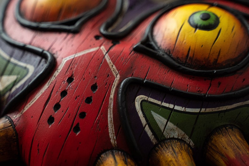 May include: Close-up of a wooden mask with a red, yellow, green, and purple paint design. The mask has a large eye with a green iris and a black pupil. The mask also has a series of small holes in the red painted area.