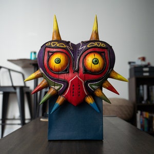 May include: A colorful, wooden mask with a heart shape and sharp points. The mask has large, yellow eyes with green pupils and a red, purple, and yellow design.