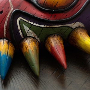 May include: Close-up of a mask with five sharp, colorful points. The points are painted in shades of yellow, green, blue, red, and brown. The mask is made of a textured material and has a wooden appearance.
