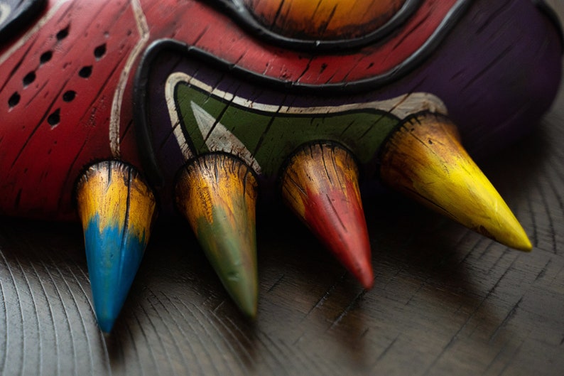 May include: Close-up of a mask with five sharp, colorful points. The points are painted in shades of yellow, green, blue, red, and brown. The mask is made of a textured material and has a wooden appearance.