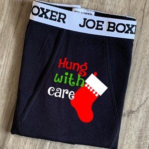 May include: Black underwear with a red and green graphic print of a stocking with the text "Hung with care". The underwear has a white waistband with the text "JOE BOXER".