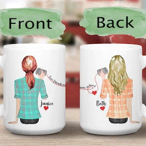 May include: Two white coffee mugs with a red string connecting them. The mugs feature illustrations of women with long hair, wearing plaid shirts, holding a can to their ear. The text on the mugs reads "True friends are never" and "...in heart". The names "Janice" and "Beth" are written on the mugs.
