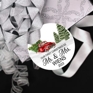 Pode incluir: Um enfeite de Natal redondo branco com um caminh&atilde;o vermelho, &aacute;rvores verdes e o texto "Our First Christmas as Mr. & Mrs. WIENS 2023".