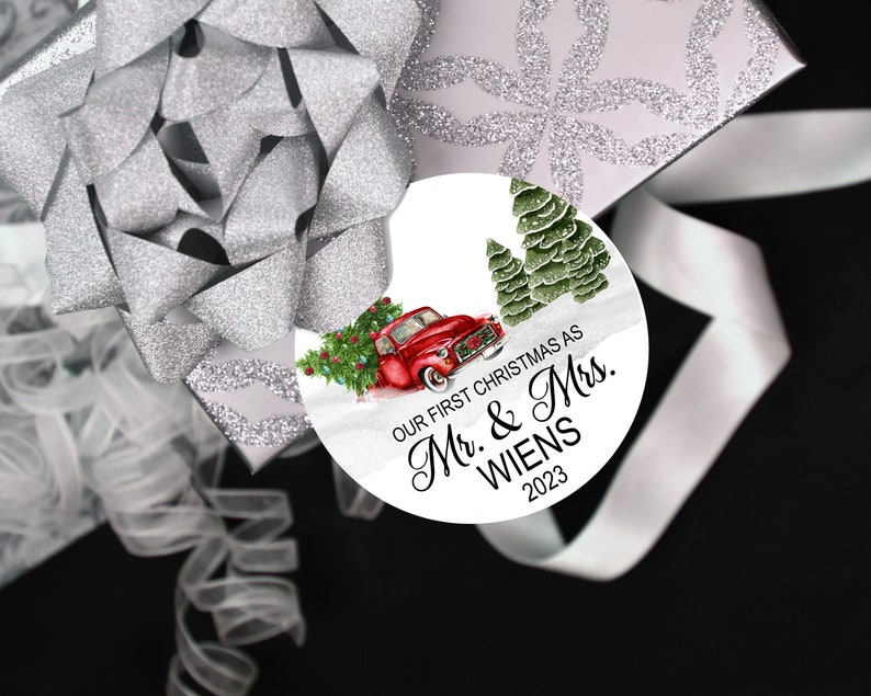 Pode incluir: Um enfeite de Natal redondo branco com um caminh&atilde;o vermelho, &aacute;rvores verdes e o texto "Our First Christmas as Mr. & Mrs. WIENS 2023".