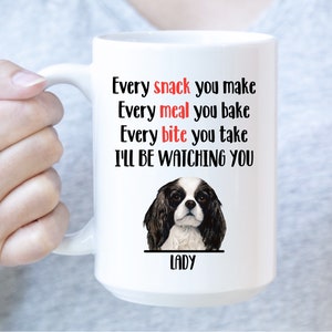 Peut inclure: Mug en céramique blanche avec le texte "Every snack you make, Every meal you bake, Every bite you take, I'LL BE WATCHING YOU." Une illustration de Cavalier King Charles Spaniel est en dessous, avec le nom "LADY" en dessous.