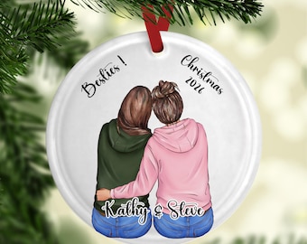 May include: A round white ceramic ornament featuring an illustration of two people from behind, wearing hoodies and jeans. The text reads Besties! Christmas 2026 and the names Kathy & Steve. A red ribbon is attached to the top for hanging.