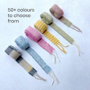 May include: Six rolls of textured fabric ribbon with frayed ends, arranged in a diagonal line. The ribbons feature various patterns and solid colours including blue plaid, light blue, pink, yellow, green, and mauve. Text reads: 50+ colours to choose from.