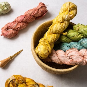 Puede incluir: Una colección de hilo de cinta de seda de colores en una cesta tejida. El hilo es en tonos de verde, amarillo, azul y rosa. También se muestra un gancho de ganchillo de madera.
