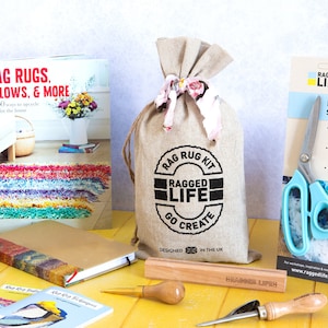 May include: A Ragged Life craft kit for making rag rugs. The kit includes a hessian bag with a printed logo, a pair of blue fabric scissors, a wooden dowel, a wooden tool, and two small books with instructions. The kit is on a yellow surface.