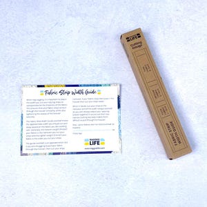 Hand using rag rug cutting gauge to cut fabric strips evenly, time-saving tool for rug hooking and peg loom weaving. Shown in cardboard packaging with fabric strip width guide