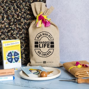 May include: A rag rug kit with a hessian bag, a small booklet, a wooden tool, a wooden block with the text "RAGGED LIFE", a blue marker with the text "RAGGED LIFE", and a small booklet with the text "Rag Rug Instructions".
