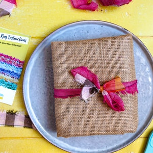 May include: A burlap wrapped gift box with a pink and orange ribbon bow. The box is sitting on a gray plate. The text on the card reads "Rag Rug Instructions - sink your toes into that!" and "Ragged Life".