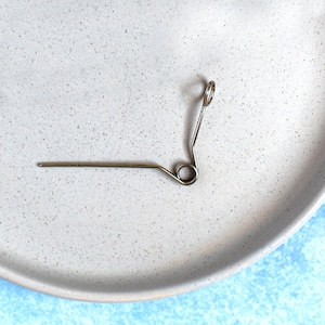 May include: A silver spring-loaded wire hook with a small loop at the end. The hook is lying on a white speckled surface.