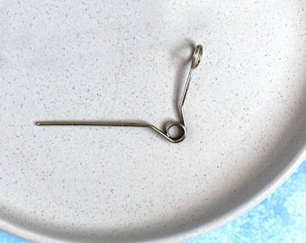May include: A silver spring-loaded wire hook with a small loop at the end. The hook is lying on a white speckled surface.