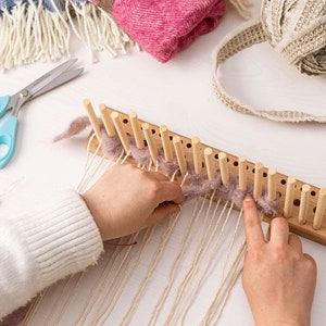 May include: A rectangular wooden knitting loom with vertical pegs, featuring light-coloured yarn being woven through the pegs. Nearby are light blue scissors, a small yellow-handled tool, a ball of beige yarn, and folded pink and blue fabric.