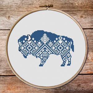 May include: A cross-stitch embroidery piece featuring a blue and white geometric pattern in the shape of a buffalo, displayed within a circular wooden hoop. The design incorporates intricate diamond and star motifs across the animal's body.