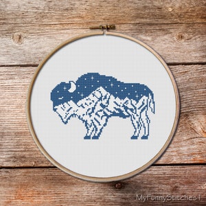 May include: A circular wooden embroidery hoop containing white fabric with a cross-stitch design of a bison. The bison is filled with a dark blue starry night sky pattern on its upper half and a white mountain range silhouette on its lower half.