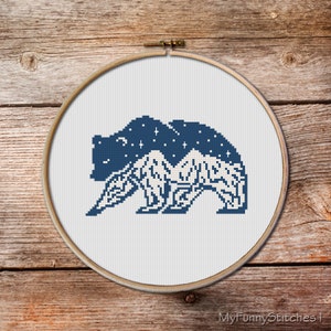 May include: A circular wooden embroidery hoop containing a cross-stitch pattern of a bear silhouette. The upper portion of the bear is dark blue with white stars, while the lower portion features a white mountain range design on a white fabric.