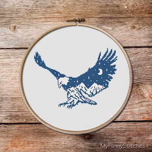 May include: A circular wooden embroidery hoop containing white fabric with a cross-stitch design of a soaring eagle in dark blue thread. The eagle's wings feature a night sky with stars and a crescent moon, while its body depicts mountain peaks.