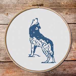 May include: A circular wooden embroidery hoop containing a cross-stitch pattern of a wolf howling at the moon. The wolf is rendered in dark blue with white star accents, while the lower portion features a white mountain range design on fabric.