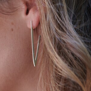 May include: Close-up of a person's ear wearing a pair of silver V-shaped earrings. The earrings are long and slender, with a minimalist design. The person has blonde hair.
