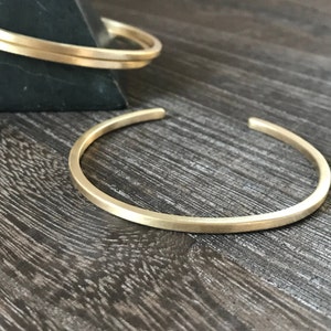 May include: Two gold-colored metal cuff bracelets. One is in the foreground, open, and in focus. The other is in the background, partially visible, resting on a dark surface. The bracelets have a simple, minimalist design.