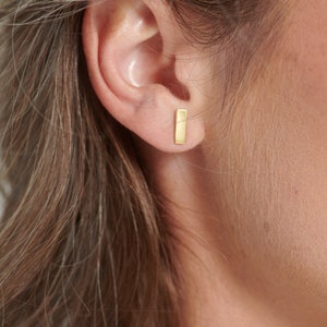 May include: A small, rectangular gold stud earring with a smooth, matte finish. The earring is positioned in the earlobe, featuring a simple, vertical bar shape that sits flush against the skin, providing a clean and understated appearance.