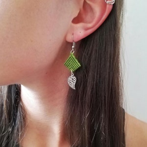 Puede incluir: Pendientes verdes y plateados con un diseño de hoja geométrica. Los pendientes están hechos de una tela tejida y presentan un delicado encanto de hoja plateada.