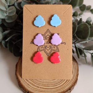 Puede incluir: Un conjunto de seis pendientes de botón de colores. Los pendientes tienen forma de nubes y corazones. Los colores son azul claro, morado y rosa.