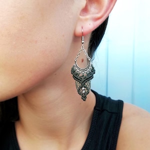 Puede incluir: Un par de pendientes de macramé verde con cuentas plateadas y un gancho plateado. Los pendientes cuelgan de la oreja de una persona.