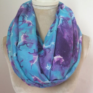 May include: An infinity scarf in shades of blue and purple, patterned with unicorns. The scarf has a celestial theme, with a light blue backdrop and purple detailing. White and purple unicorns are featured.
