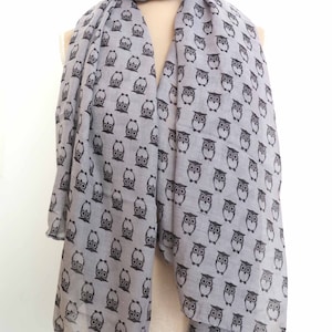 May include: A pale grey scarf featuring a repeating pattern of black owl motifs. The scarf is displayed on a mannequin, highlighting its drape and design. The owls have large eyes and are in various poses.
