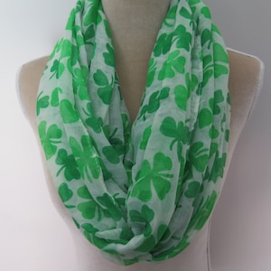 May include: An infinity scarf in green and white, patterned with shamrocks. The scarf is made of a light fabric and is displayed on a mannequin. The shamrocks are a vibrant green colour, perfect for St. Patrick's Day.