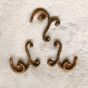 May include: Three decorative metal hooks with a bronze finish. Each hook has a scrolled design, with a curved top and a downward-facing hook. The hooks are arranged on a textured, off-white surface, suitable for a vintage aesthetic.