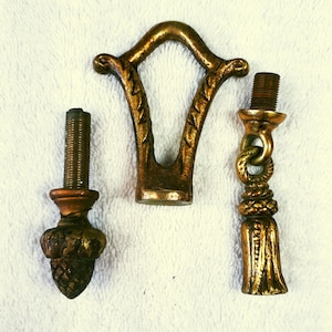 May include: Three antique brass lamp components. These include a decorative finial, a V-shaped bracket, and a tassel-shaped piece. The finial has a threaded base and an acorn design. The bracket has a decorative leaf pattern.