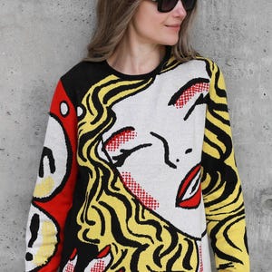 May include: A black, yellow, red, and white pop art style sweater featuring a woman's face with a red mouth and yellow hair.