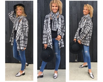 May include: A woman wearing a black and white patterned cardigan, a black scarf, blue jeans, and silver loafers. She is holding a black hat in her hand.