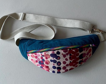May include: A teal blue corduroy fanny pack with a pink and white patterned lining. The fanny pack has a rainbow zipper and a white adjustable strap.
