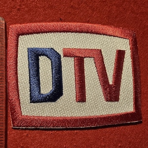May include: Embroidered patch with the letters "DTV" in blue and red on a white background, framed by a red border. The patch is approximately 3 inches long.