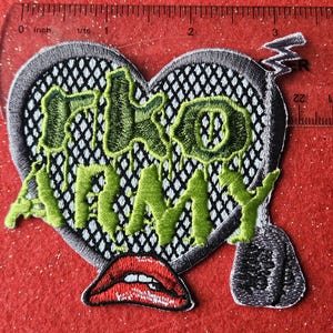 May include: Embroidered patch in the shape of a heart with the words "FKO ARMY" in green, dripping letters. The heart has a black mesh design and a gray border. A red lip graphic is at the bottom.