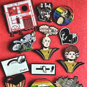 May include: A collection of twelve enamel pins featuring various designs including a red and white laboratory machine, a group of people, a rainbow circle, a motorcycle with the name Eddie, a person with a yellow shirt, a heart, and lips.