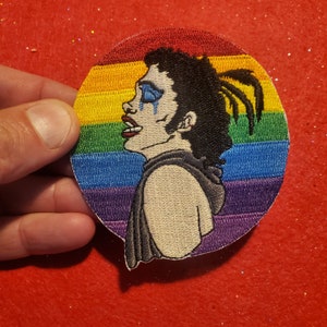 以下が含まれることがあります： 虹色の刺繍パッチ。青いアイシャドウと頬を伝う涙をした人物の横顔が描かれています。人物は黒い衣服を着ており、虹色の背景に配置されています。