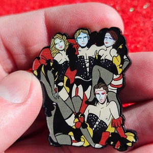 May include: A black enamel pin featuring a group of four figures in a cabaret setting. The figures are wearing colorful costumes and makeup, with one figure in a red and black costume and another in a yellow and black costume. The pin is a stylized representation of a cabaret scene.