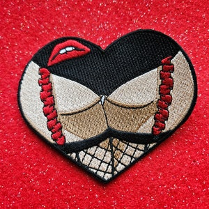 Puede incluir: Un parche bordado en forma de corazón con fondo negro y un diseño de sujetador beige. El sujetador tiene tirantes rojos y un diseño de labios rojos en la parte superior.