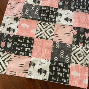 May include: A pink and black patchwork baby blanket with a buffalo plaid pattern, arrows, and the words "Wild Wild" repeated throughout.