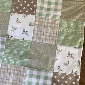 May include: A patchwork quilt featuring squares of various patterns and colors. The quilt includes squares with duck illustrations, green gingham, and geometric designs in shades of brown, green, and white. The quilt is likely made of cotton.