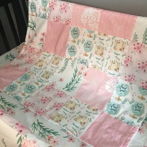 May include: A pink and white patchwork baby blanket with a travel theme. The blanket features a map, globes, suitcases, and floral patterns. The text "Travel the world" is repeated throughout the blanket.