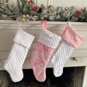 May include: Three Christmas stockings hanging on a white mantel. One stocking is white with white polka dots, one is pink with white polka dots, and one is pink with a fuzzy texture.