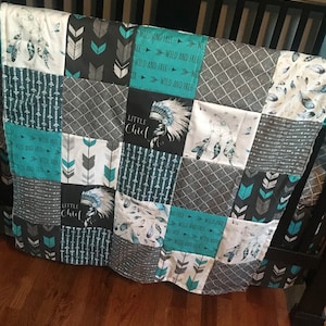 May include: A turquoise and gray baby quilt with a patchwork design. The quilt features a variety of patterns, including arrows, feathers, and dreamcatchers. The quilt also includes the text "Little Chief" in a black font.