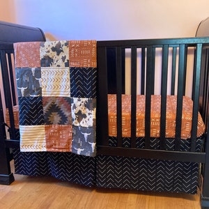 May include: A black wooden crib with a brown and black patterned crib sheet and a patchwork quilt with a cow print, geometric patterns, and a brown and white skull design.